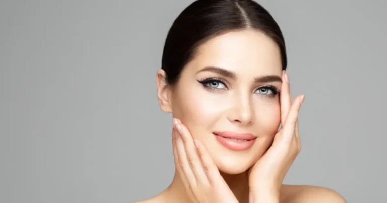 Close-up of a smiling woman with clear, smooth skin gently touching her face against a clean grey background.
