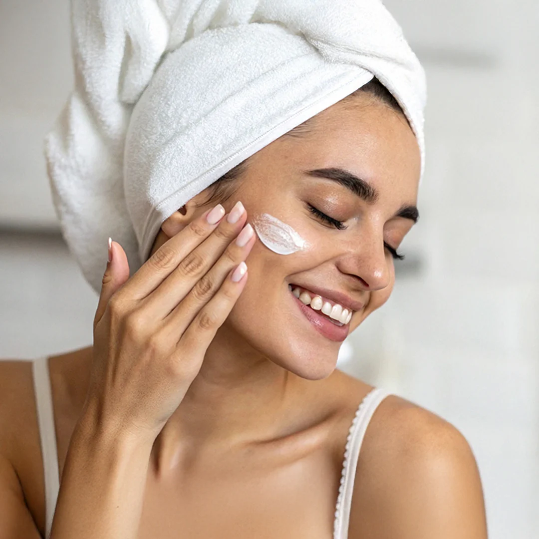 A smiling woman with a towel wrapped around her head gently applies white face cream to her cheek, wearing a light tank top and standing in a softly lit indoor setting.